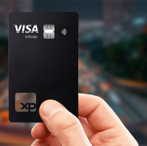 Mão segurando um cartão preto fosco da XP Visa Infinite com chip metálico e símbolo de pagamento por aproximação, tendo ao fundo uma cidade iluminada à noite.
