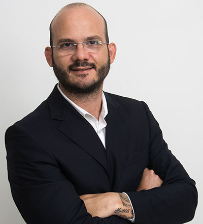 Fotografia em estúdio de meio corpo do professor e advogado Luciano Gagno. Ele é um homem branco de meia-idade, calvo, com barba cheia e bem cuidada, e usa óculos de grau com armação metálica fina. Veste um terno azul-marinho alinhado sobre camisa social branca com o colarinho aberto. Está com os braços cruzados, sorri de forma sutil e confiante para a câmera, revelando uma tatuagem escura no pulso esquerdo. O fundo é neutro em tom claro.