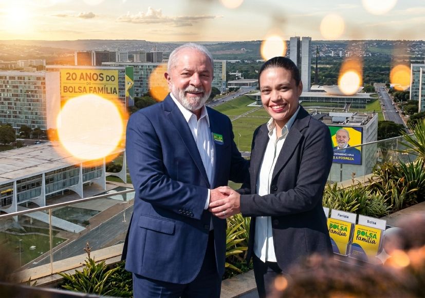 A candidata a deputada federal Jaqueline Lopes, de blazer preto, sorri ao apertar a mão do Presidente Lula, que veste terno azul, em uma varanda com a Esplanada dos Ministérios ao fundo.