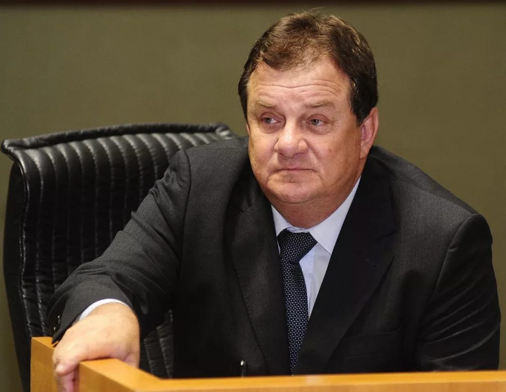 O ex-deputado e ex-prefeito José Carlos Elias, vestindo terno preto e gravata de bolinhas, sentado em uma cadeira de couro com o braço sobre um púlpito de madeira.