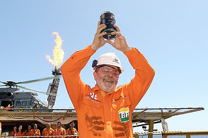Jornal o Centro – Espírito Santo: Pré-Sal: riqueza, educação e futuro Presidente Luiz Inácio Lula da Silva, vestindo macacão laranja e capacete branco, segura amostra de petróleo do pré-sal durante cerimônia no campo de Jubarte, litoral do Espírito Santo, com plataforma e helicóptero ao fundo